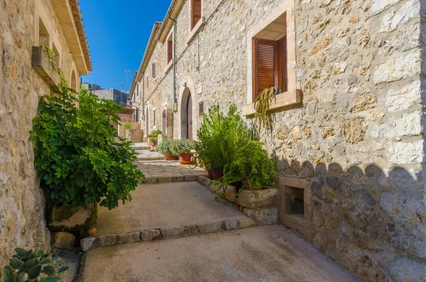 A sunlit stone alley runs between buildings, with potted greenery along the walkway.