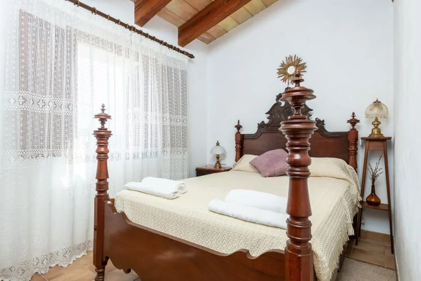 A bedroom with a wooden bed, folded towels, and lace curtains.