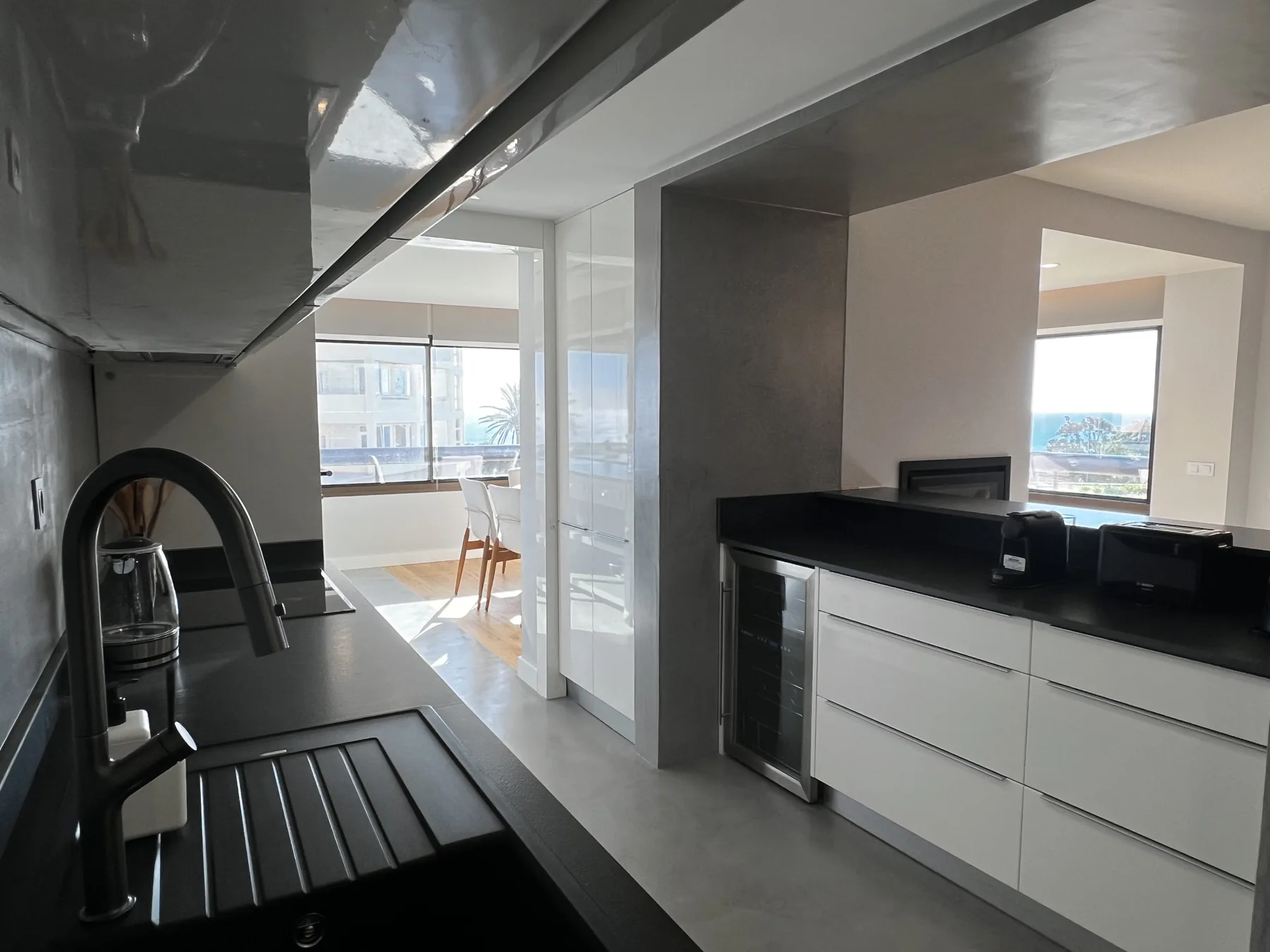 A modern kitchen opens onto a dining area beside large windows.