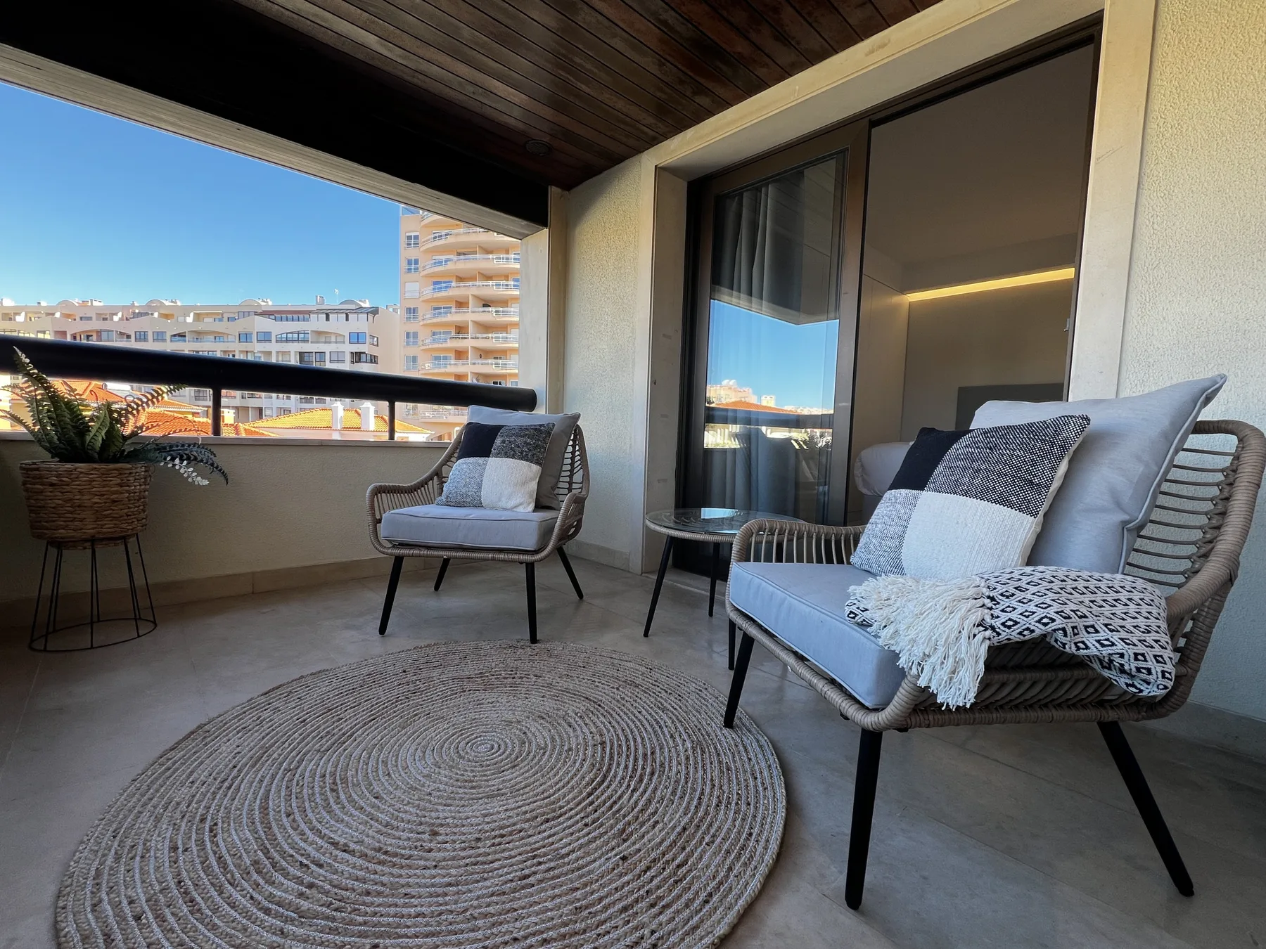 A covered balcony seating area with a round woven rug and views of nearby buildings.
