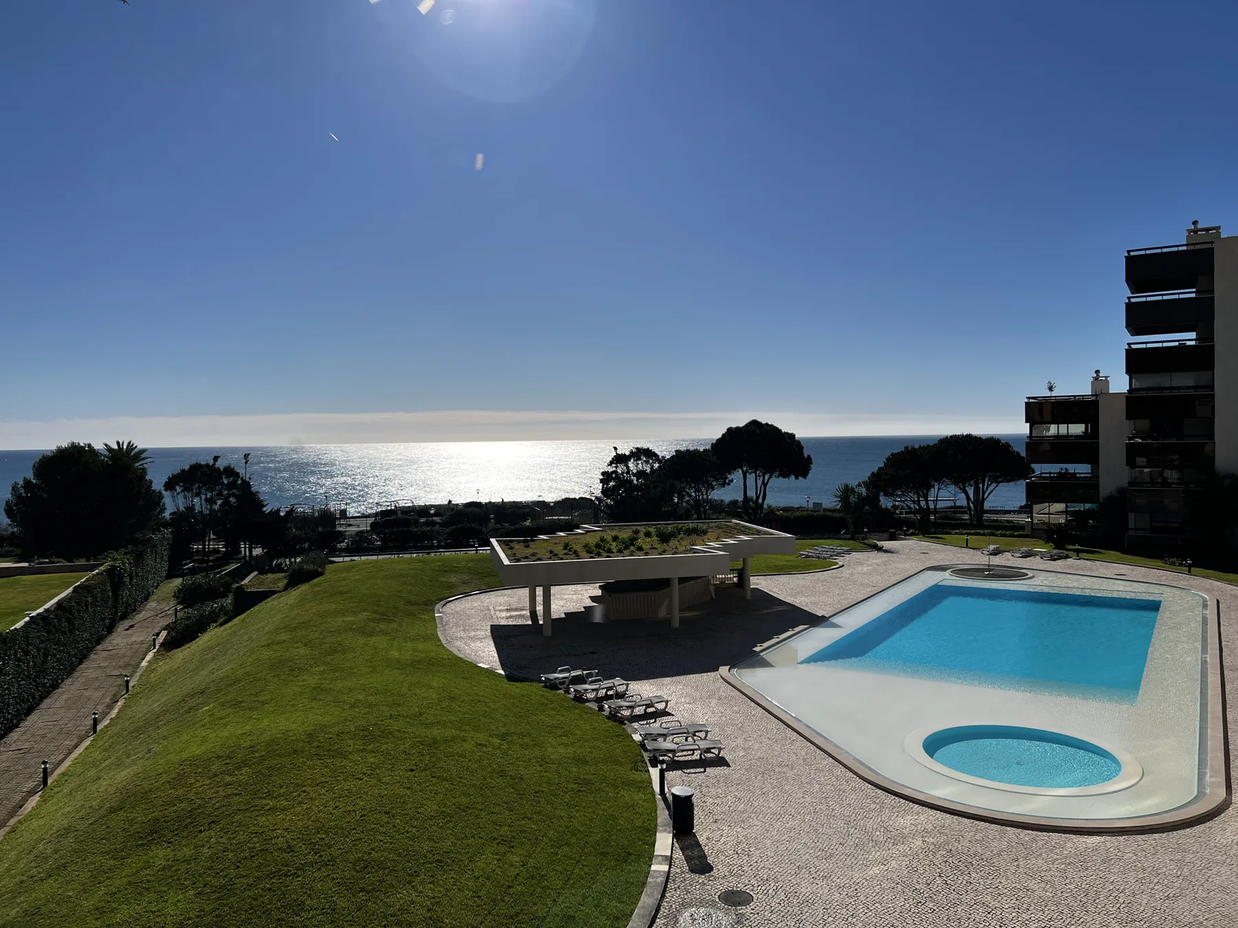 A swimming pool and landscaped grounds overlook the ocean beneath a clear sky.