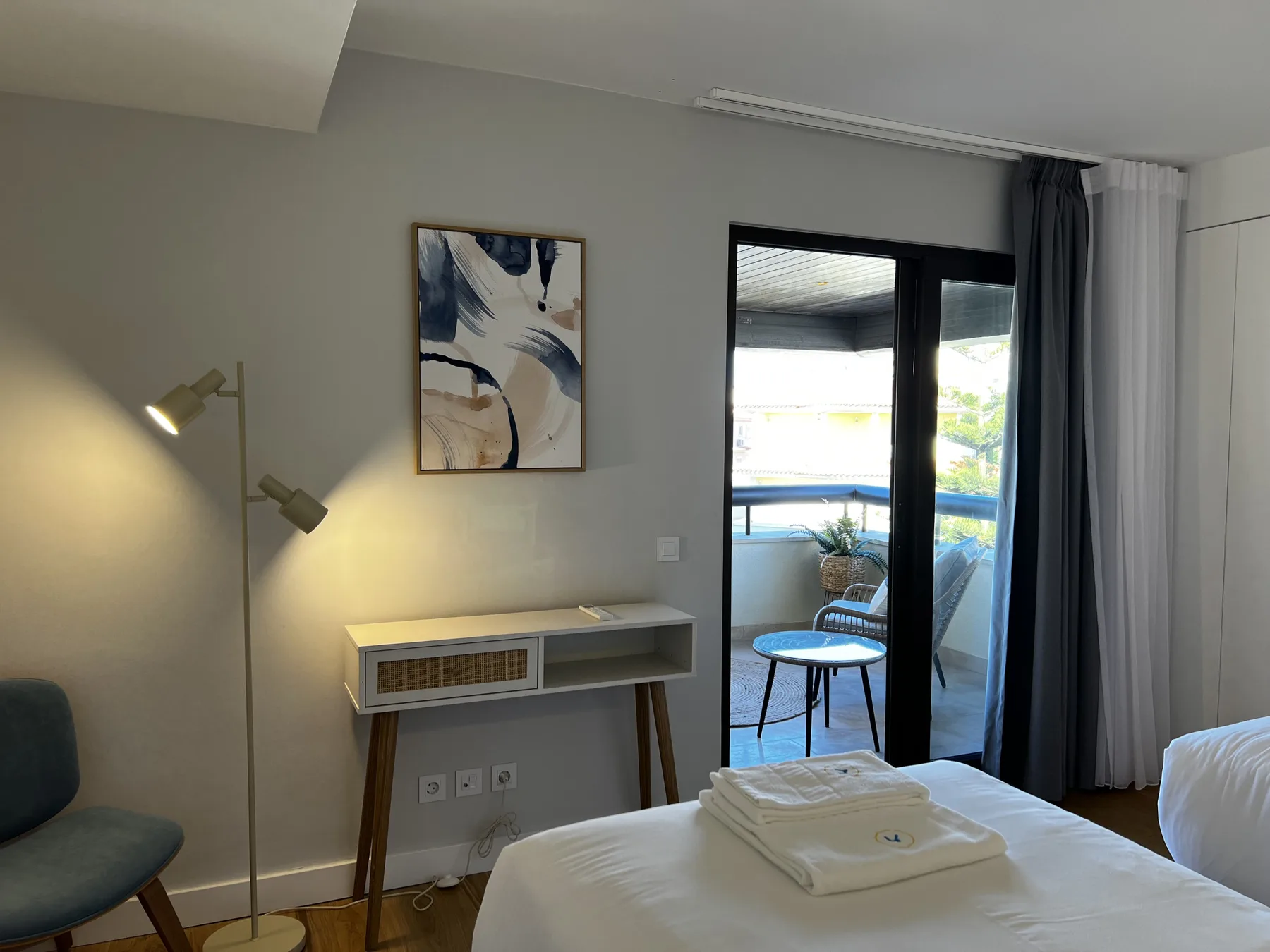 A bedroom with a bed, a small table, and an open glass door to a balcony.