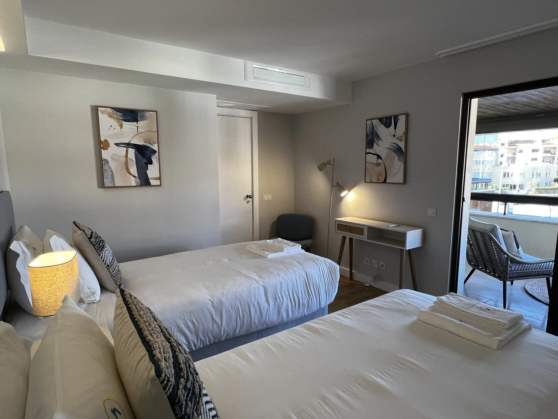 A bedroom with two beds, bedside lamps, and a desk beside a balcony.