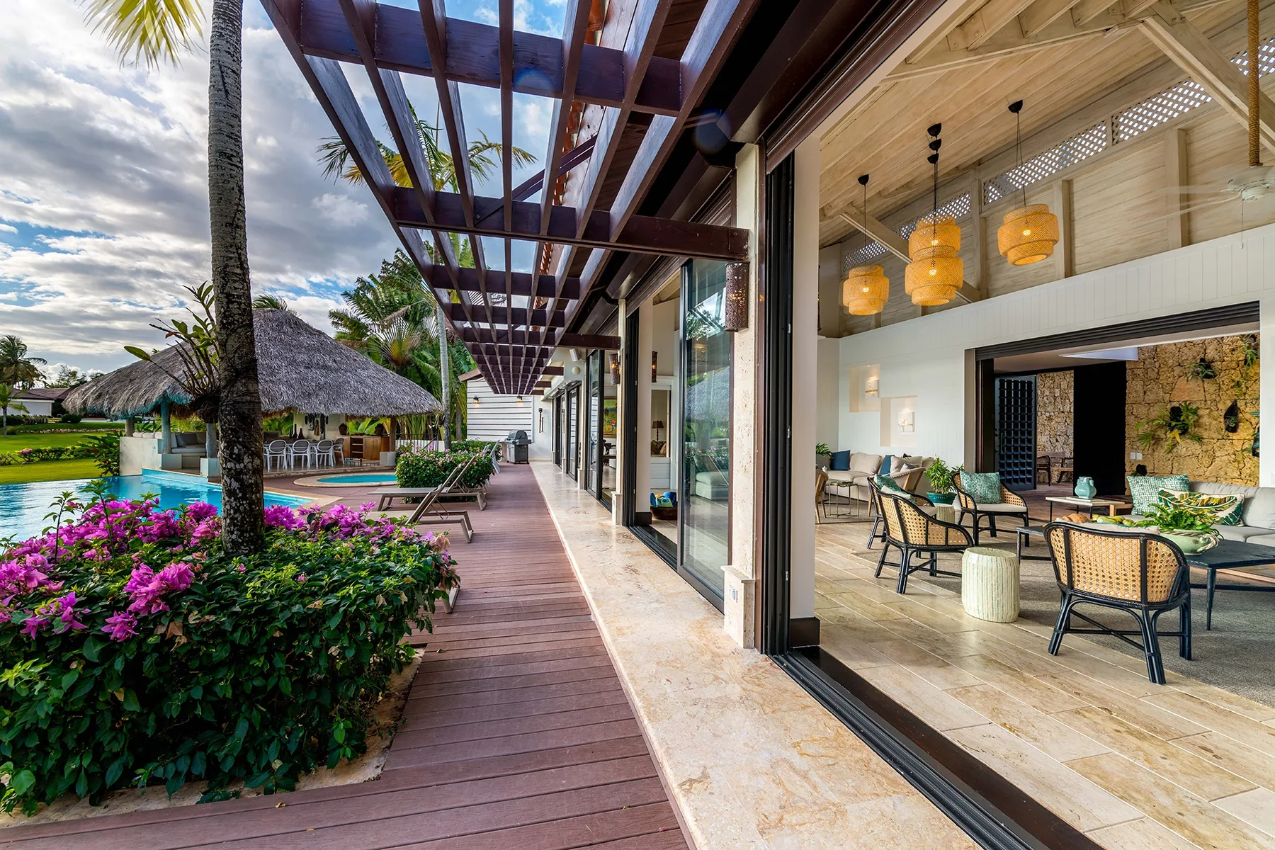 Una terraza exterior cubierta junto a un comedor, con una piscina y plantas tropicales cerca.