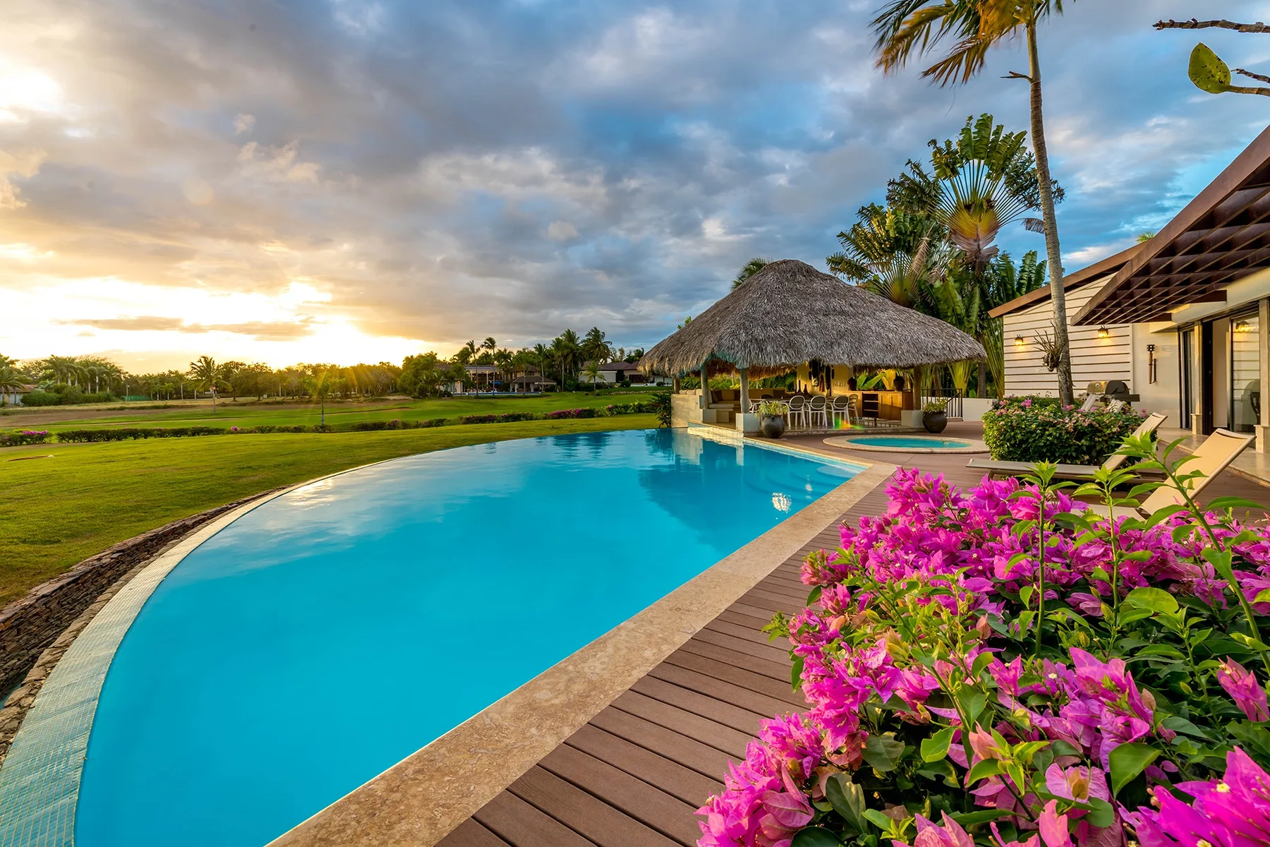 Una piscina curva junto a una terraza de madera, plantas tropicales y un pabellón con techo de paja.