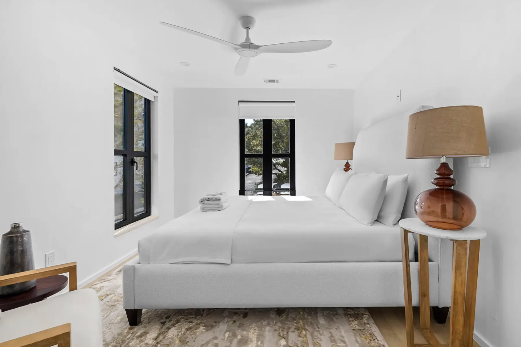 Minimalist bedroom with a large bed, white bedding, two wooden lamps, a ceiling fan, and windows with natural light. Neutral decor and carpet.