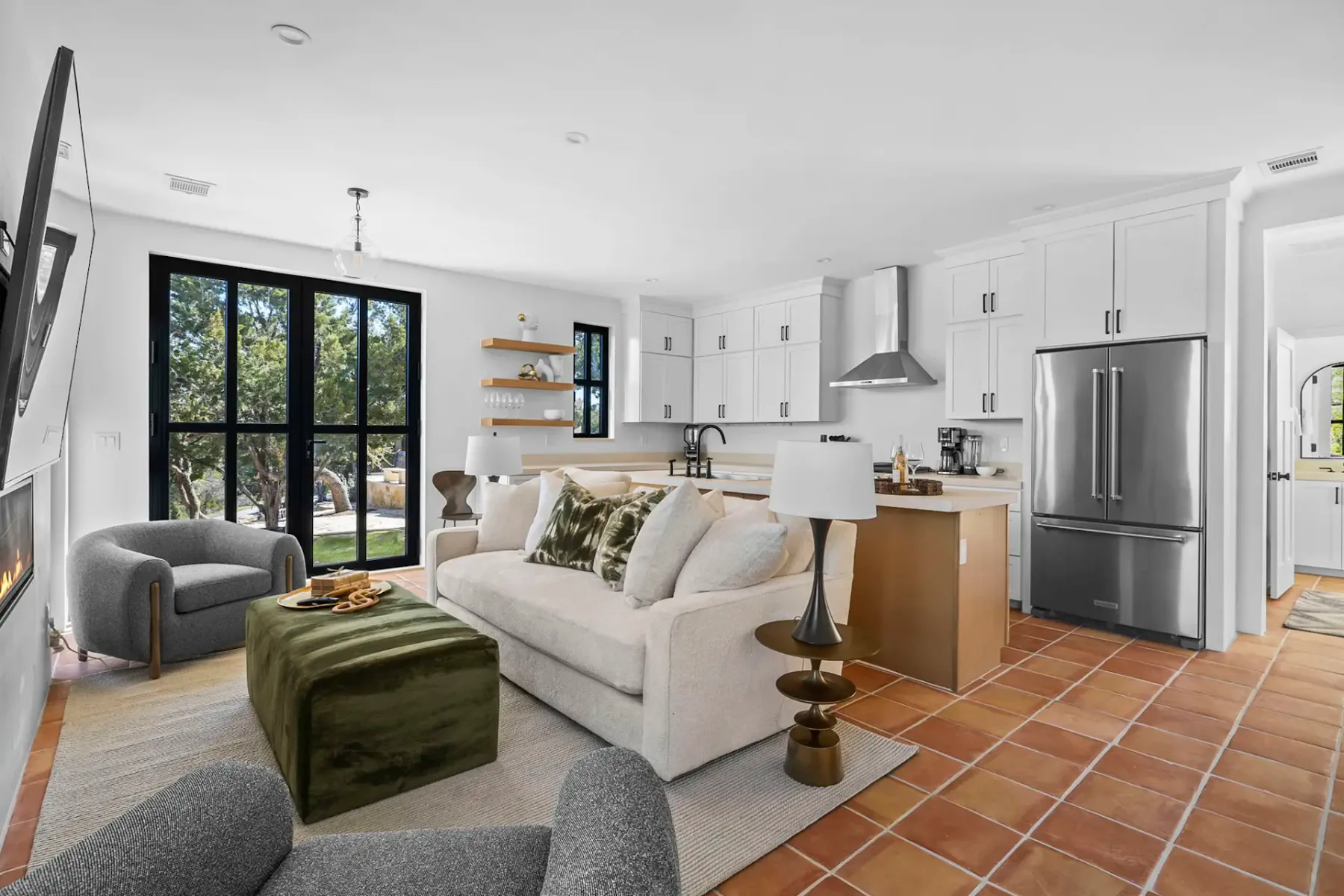 Modern open-plan living room and kitchen with white cabinets, stainless steel appliances, a cozy sofa, armchairs, and large windows overlooking greenery.