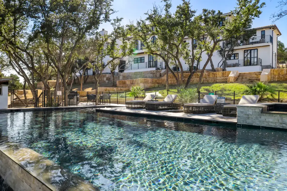 Outdoor pool with clear water, surrounded by lounge chairs and trees, facing a large, white two-story building in the background.