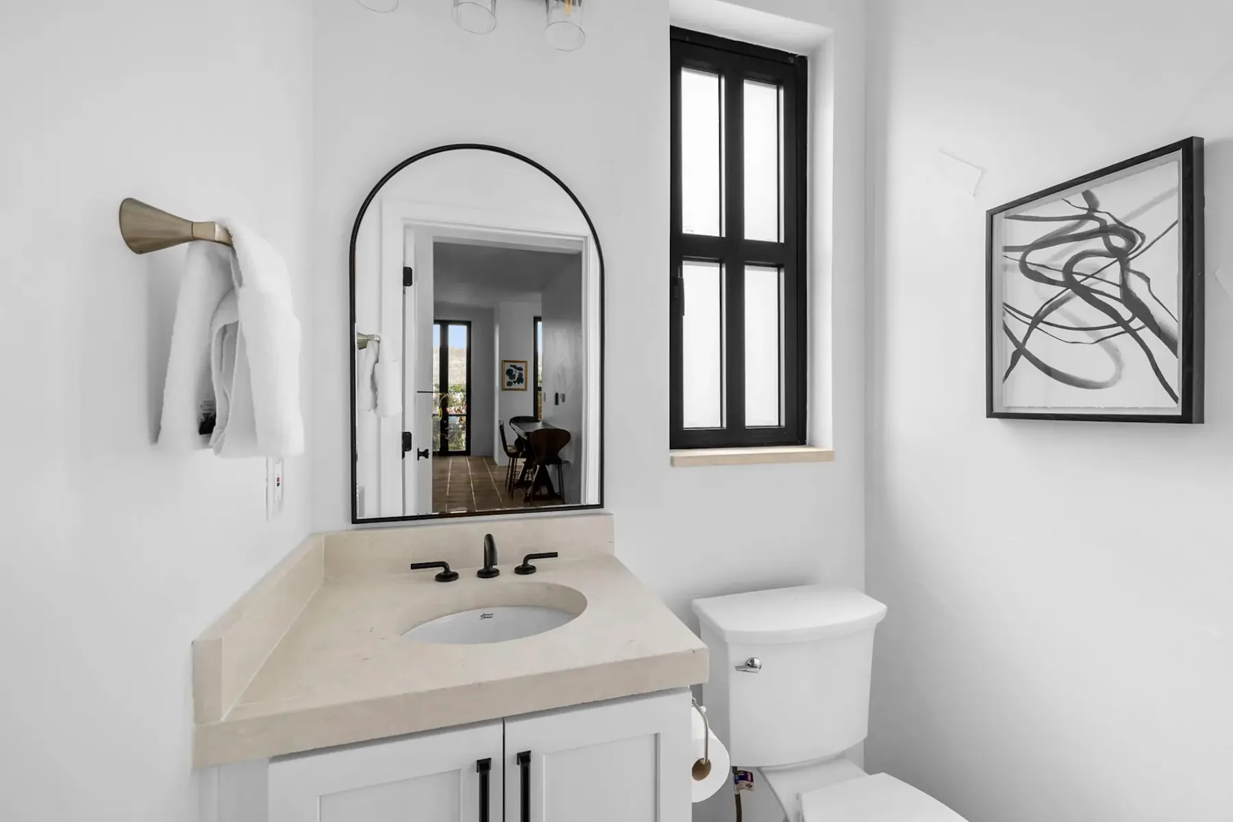 Modern bathroom with a beige countertop, black fixtures, an arched mirror, abstract wall art, towel, and window with black frame.