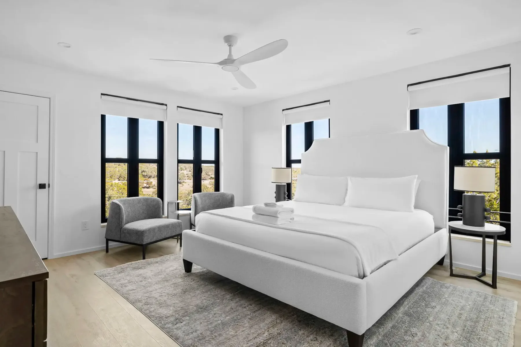 Bright bedroom with white bed and linens, two gray chairs by large windows, a dark dresser, and a ceiling fan, on light wooden flooring.