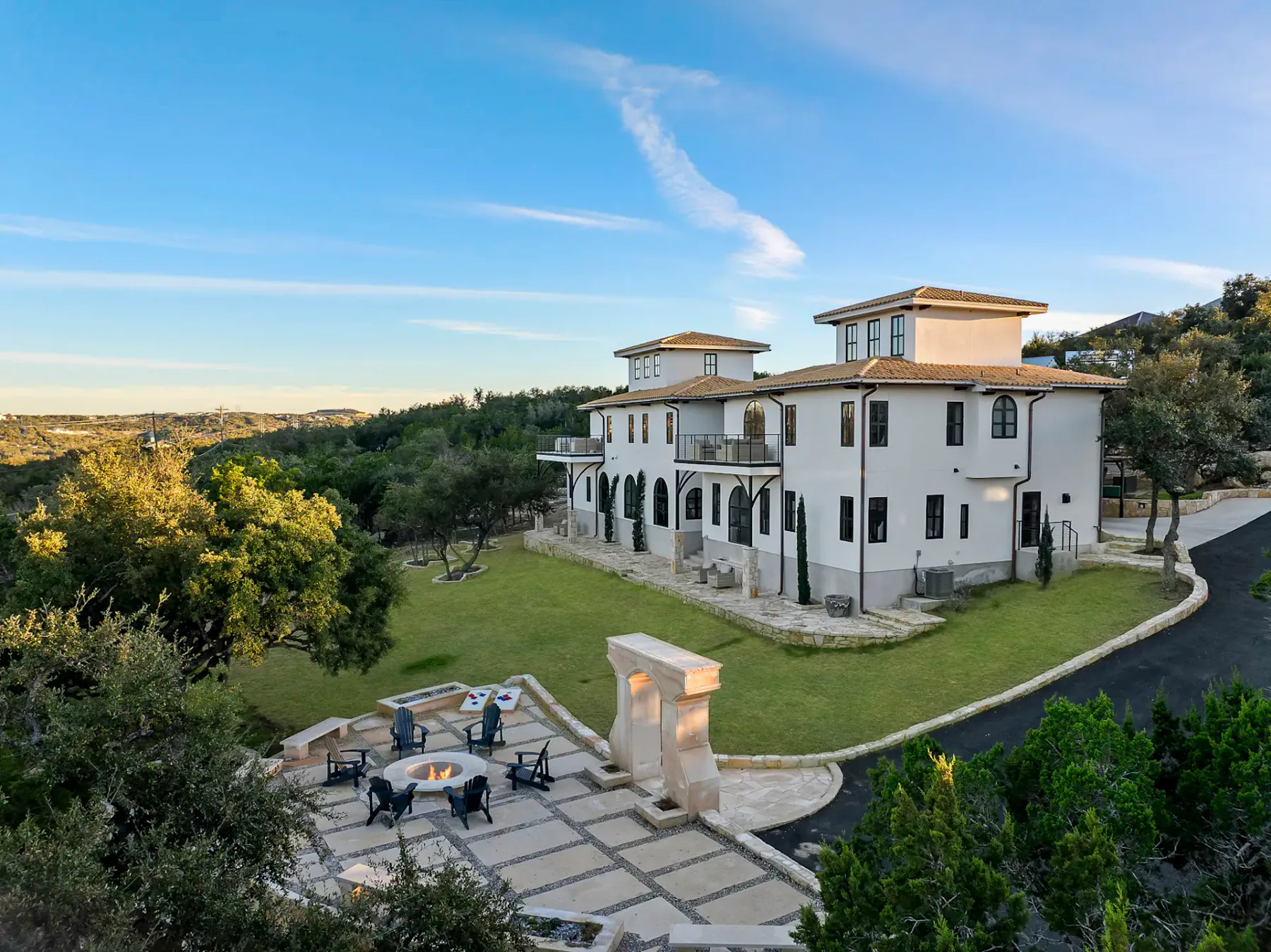 A large white Mediterranean-style villa with two prominent towers, surrounded by trees, a spacious lawn, and a paved patio with outdoor seating.