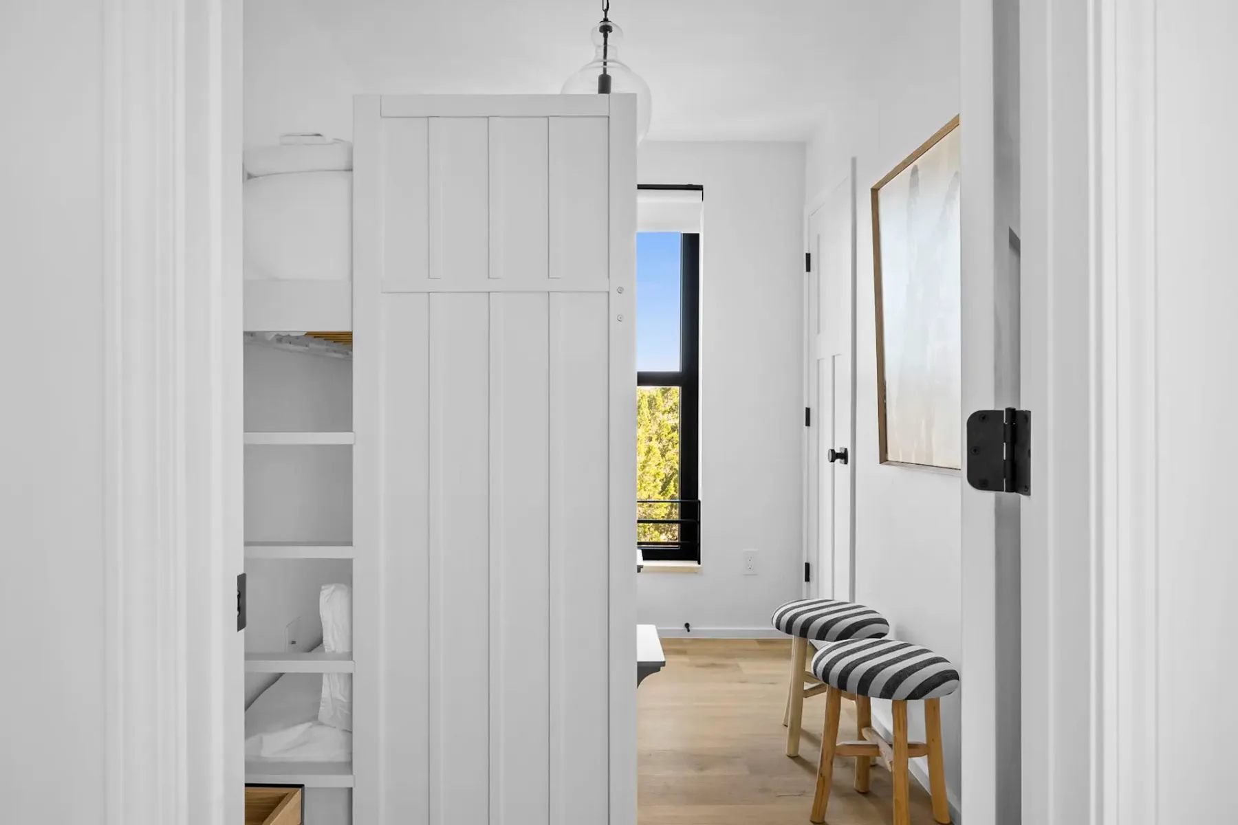 A bright bedroom with a partly open bunk bed and striped stools beside a tall window.