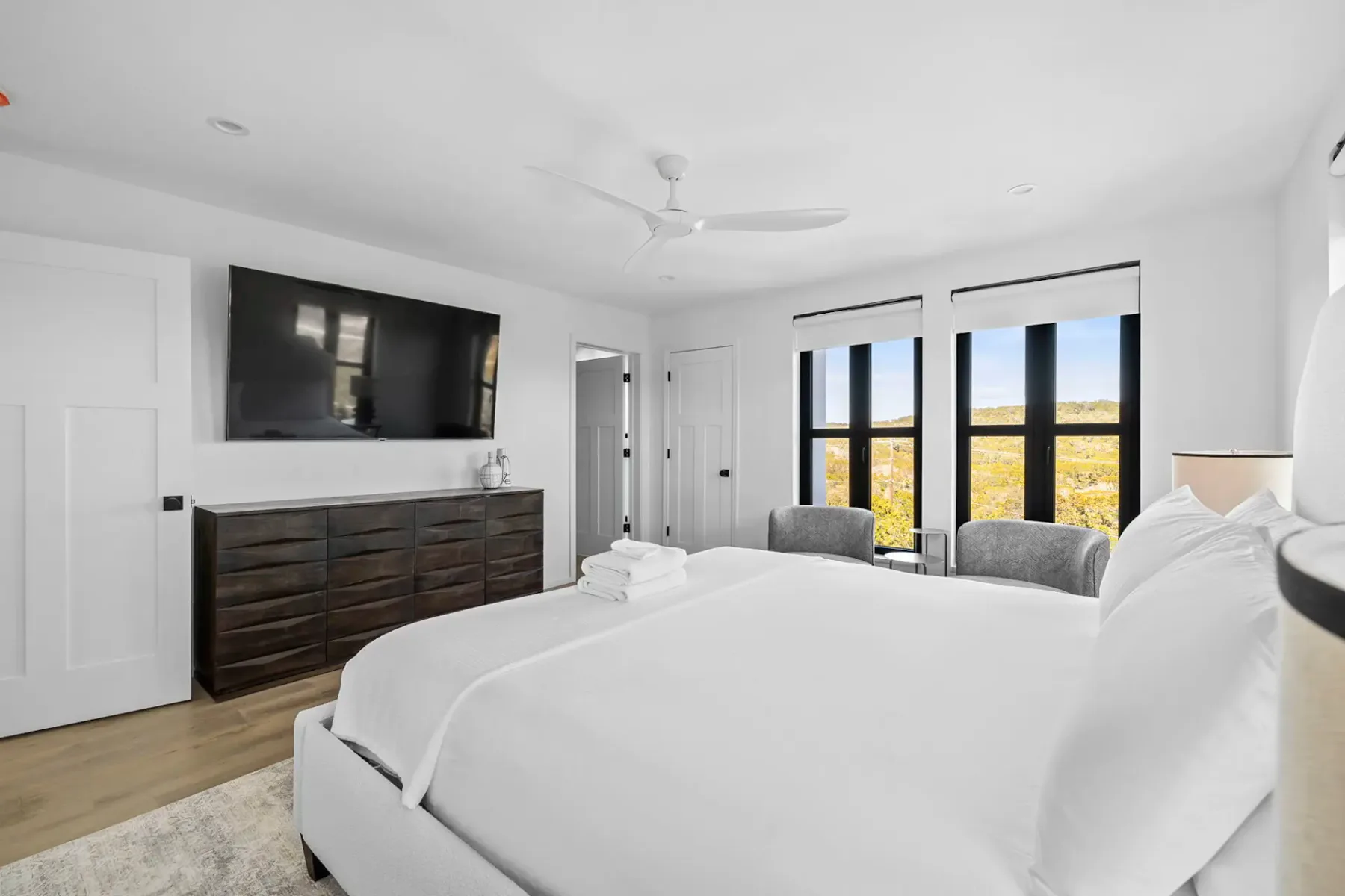 Bright bedroom with white bed and linens, two gray chairs by large windows, a dark dresser, and a ceiling fan, on light wooden flooring.