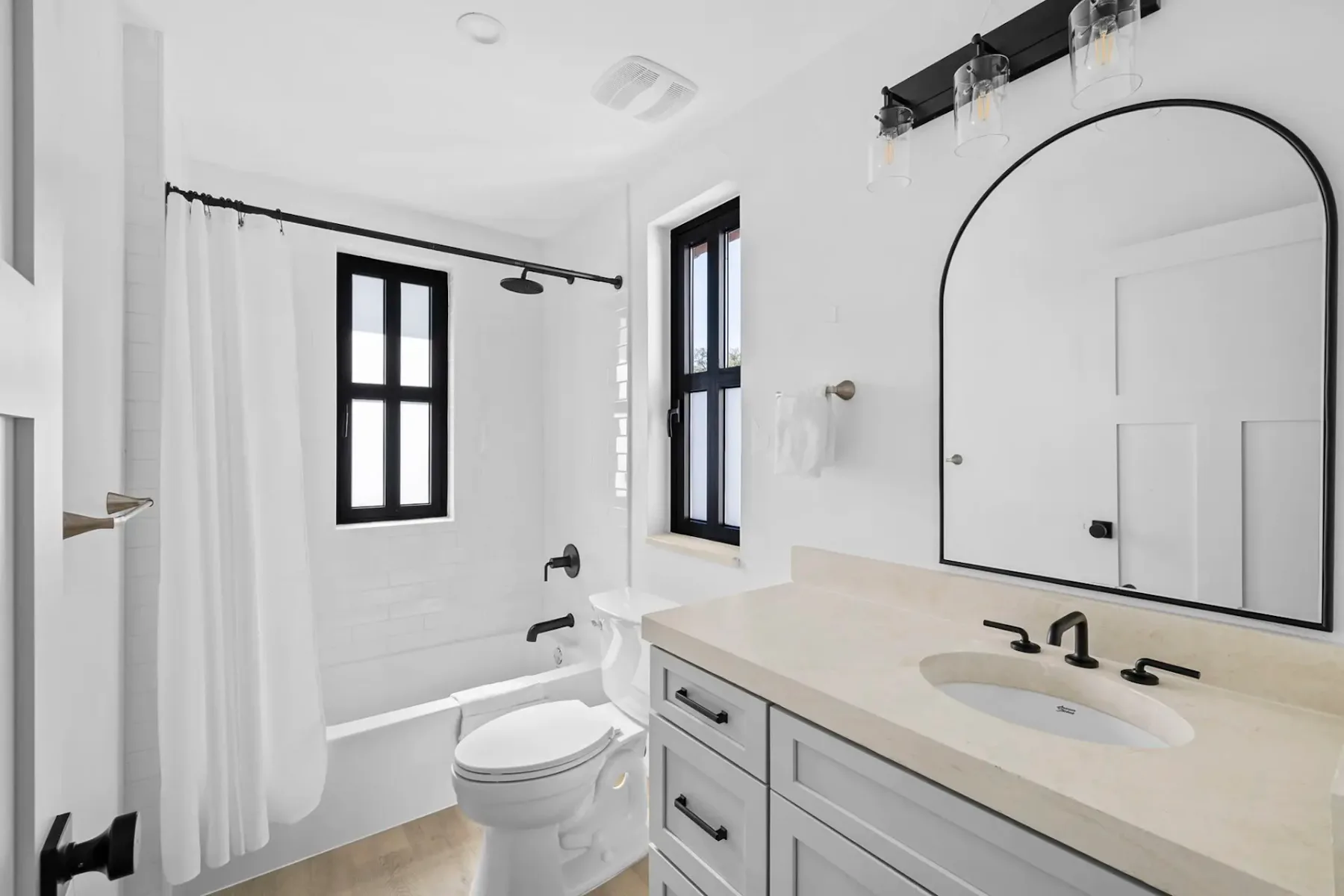 A modern white bathroom with a bathtub, black fixtures, a rounded mirror above a sink, and windows letting in natural light.