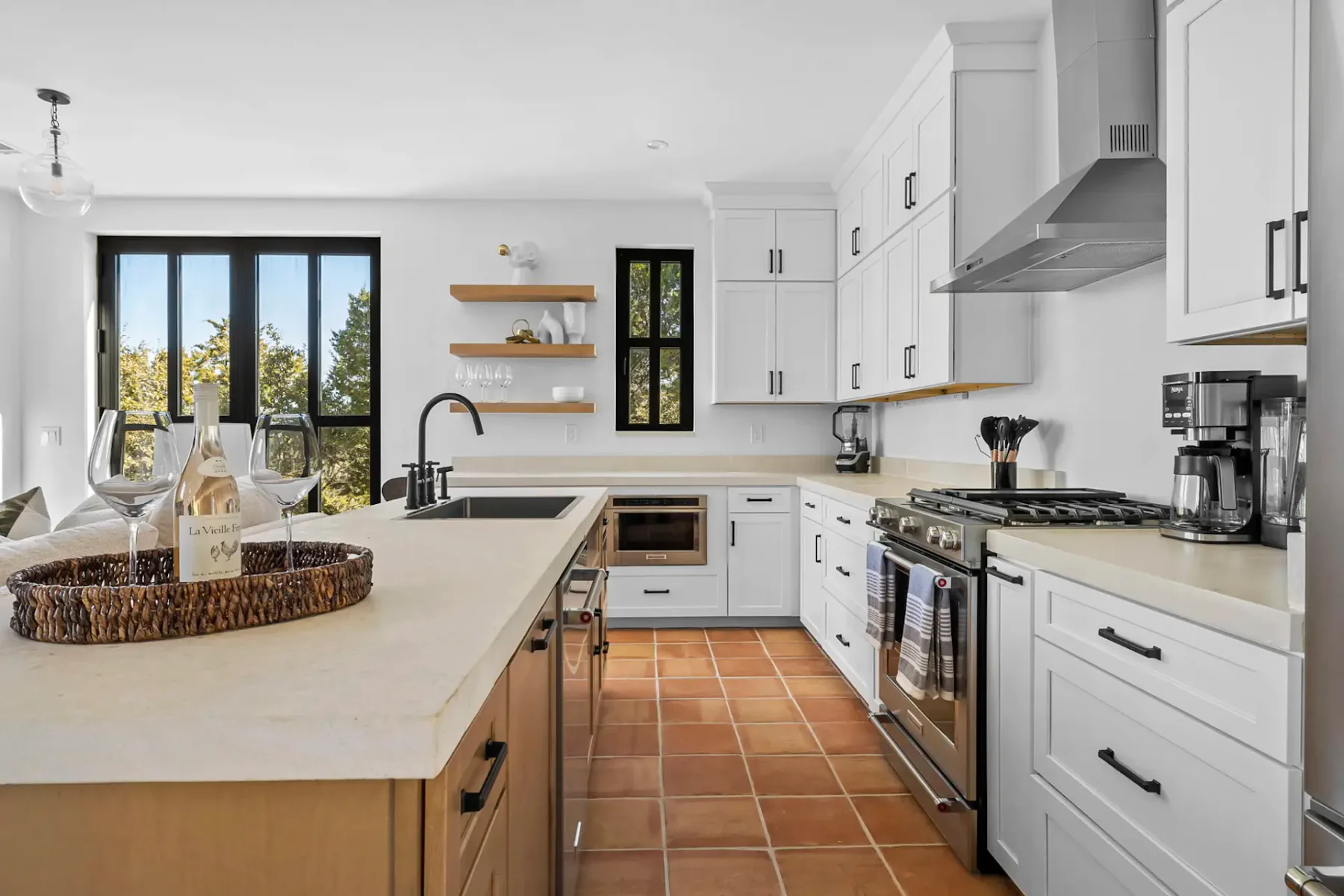 Modern kitchen with white cabinets, stainless steel appliances, a central island with a tray of glasses, and terracotta floor tiles.