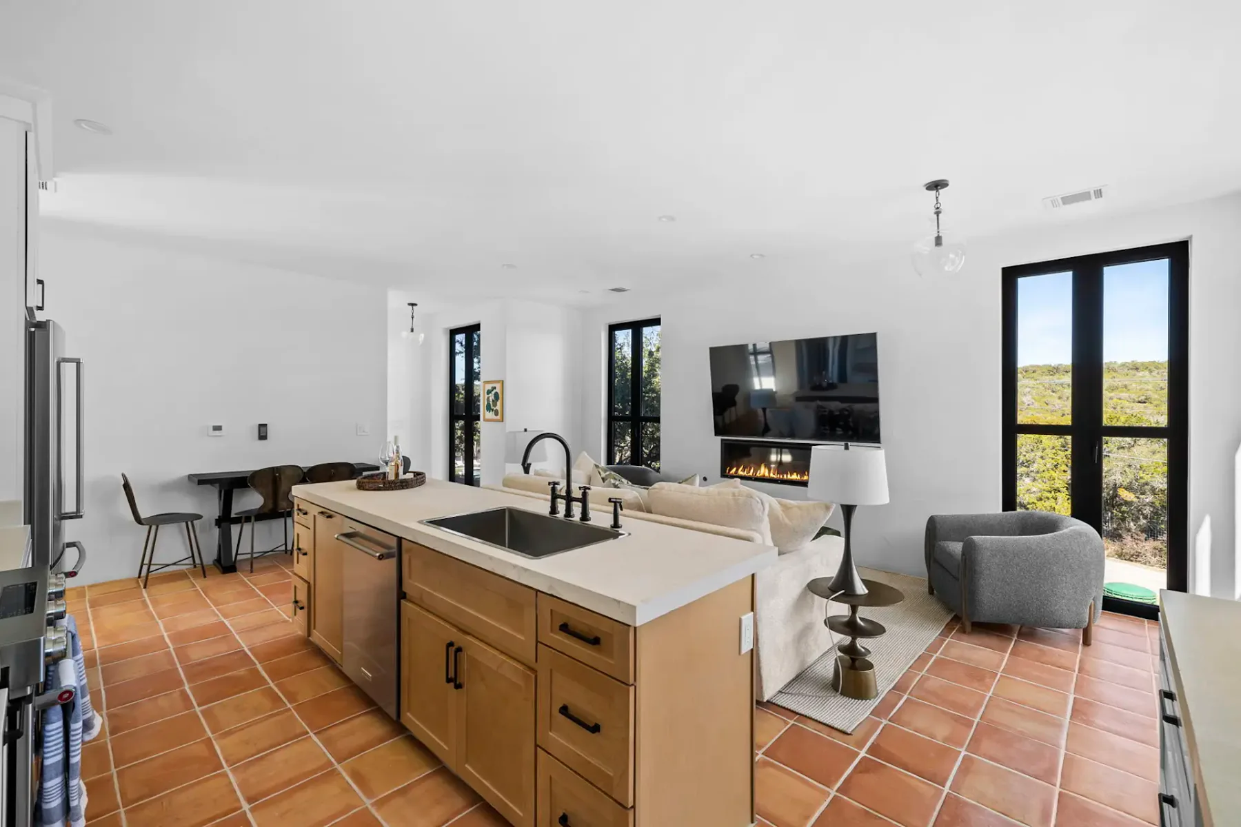 Open-plan living area with terracotta tiled floors, a kitchen island, seating area with a sofa, and a wall-mounted TV, lit by natural light.