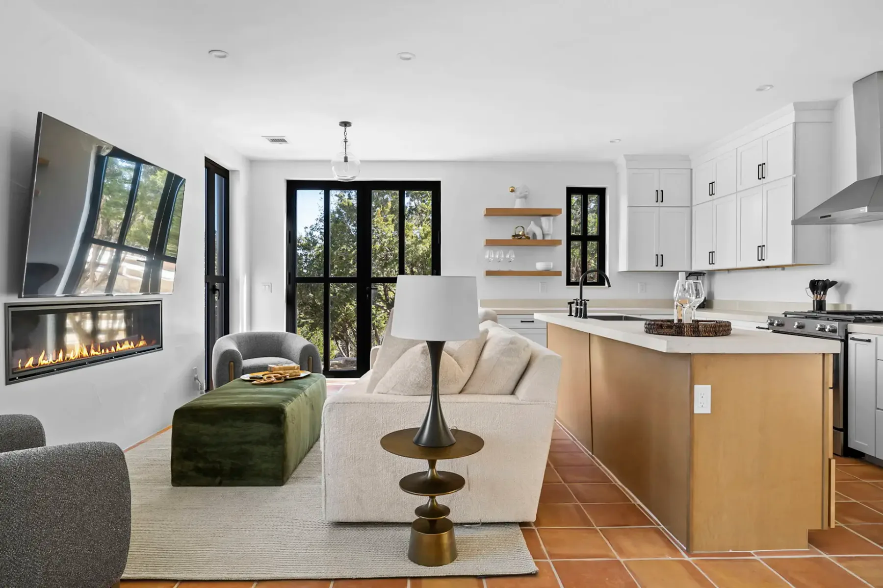 Modern open-plan living room and kitchen with white cabinets, stainless steel appliances, a cozy sofa, armchairs, and large windows overlooking greenery.