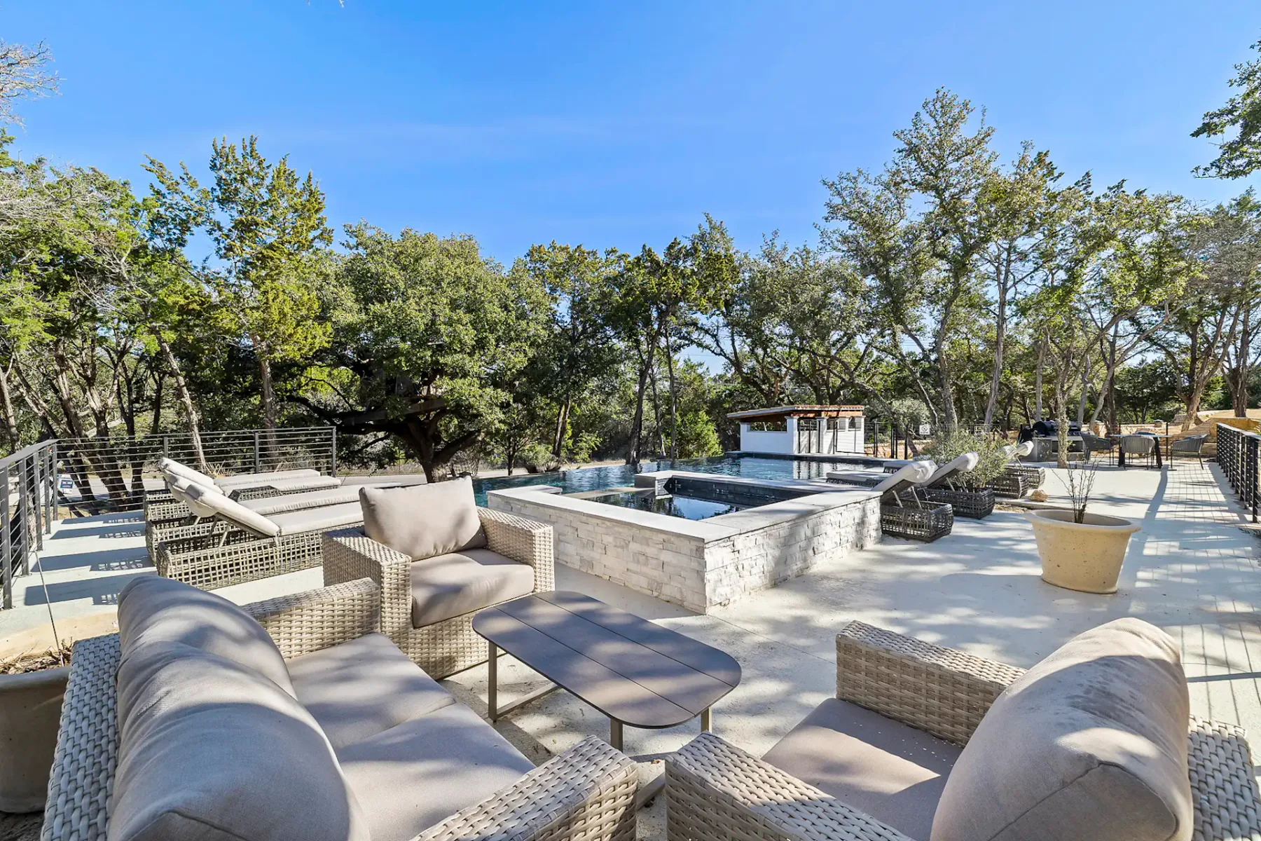 Outdoor patio with wicker sofas, a rectangular pool, and surrounded by trees. Clear blue sky and modern decor enhance the serene setting.