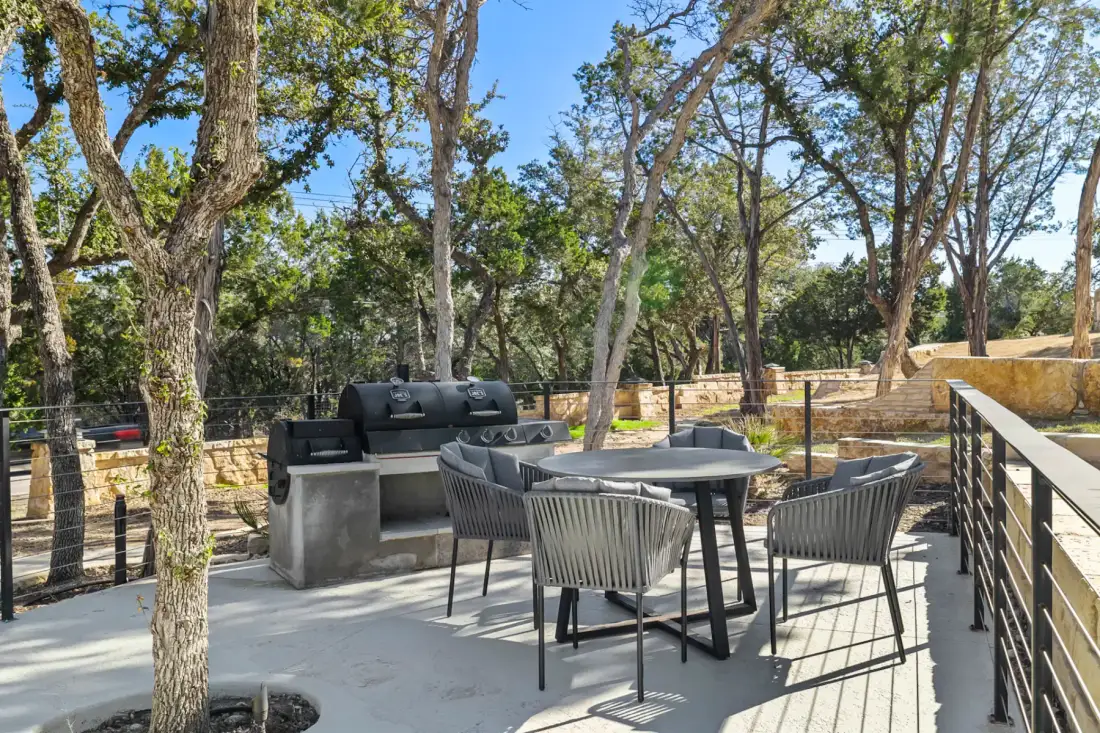 Outdoor patio with a black grill, round table, and chairs surrounded by trees and a stone fence under a clear blue sky.
