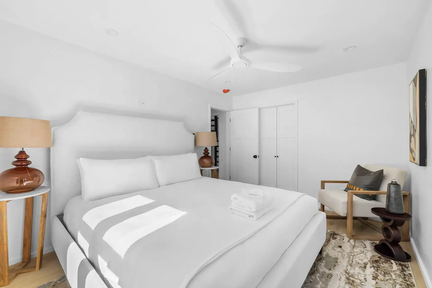 Minimalist bedroom with a large bed, white bedding, two wooden lamps, a ceiling fan, and windows with natural light. Neutral decor and carpet.