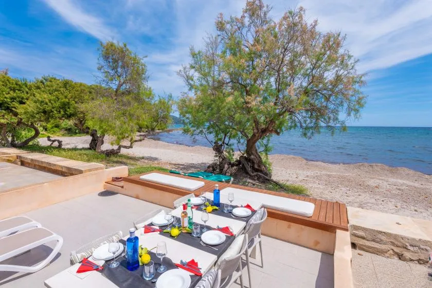 A seaside patio has a dining table set beside the beach and water.
