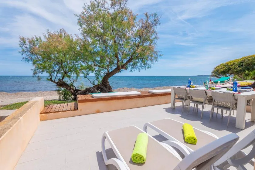 A seaside patio with loungers, dining seating, and a tree overlooking the water.