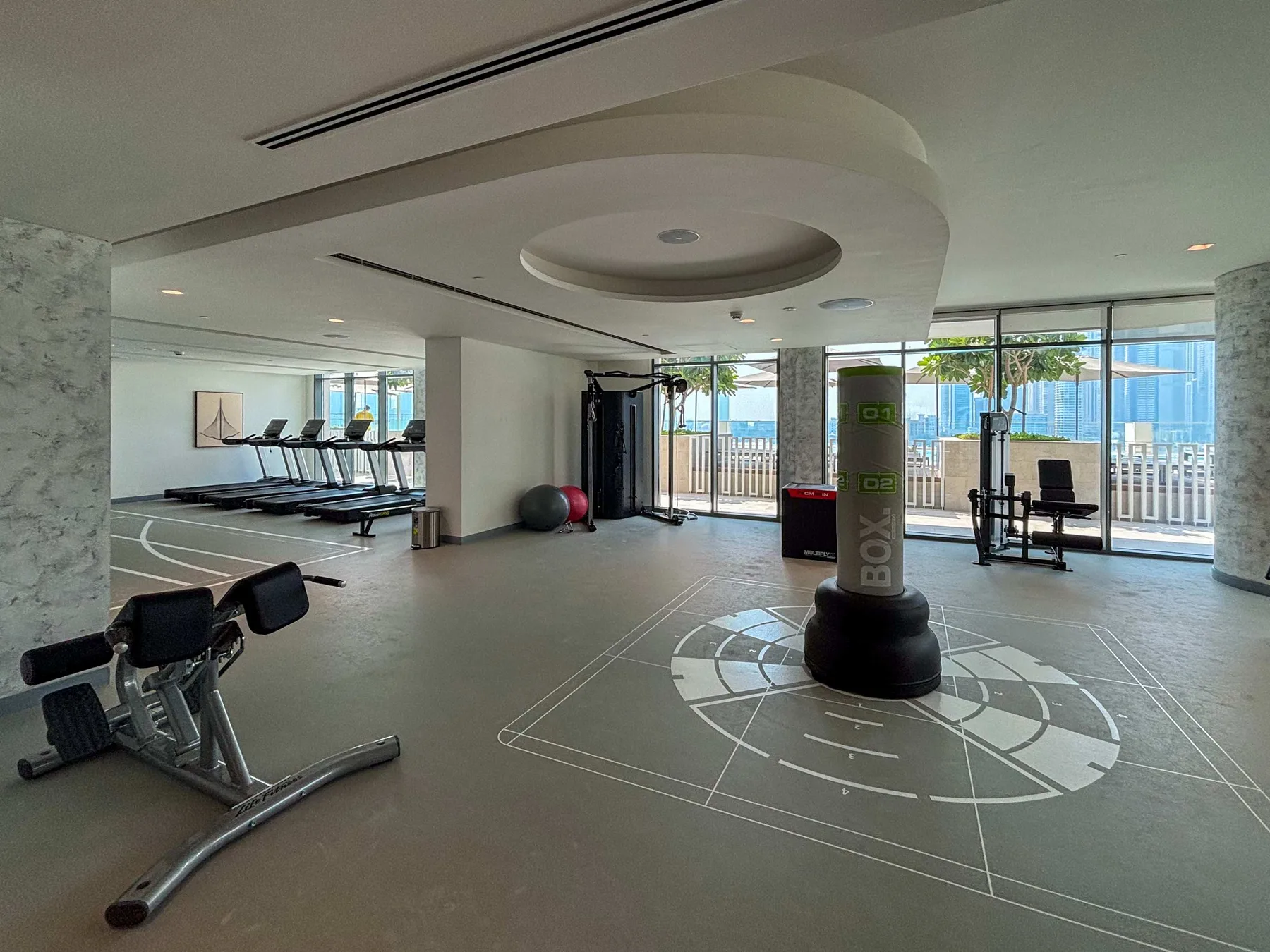 Una sala de ejercicio luminosa con equipos de entrenamiento y grandes ventanales al exterior.