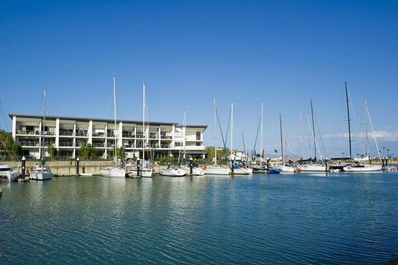 Un puerto deportivo con veleros está junto a un edificio frente al agua bajo un cielo despejado.