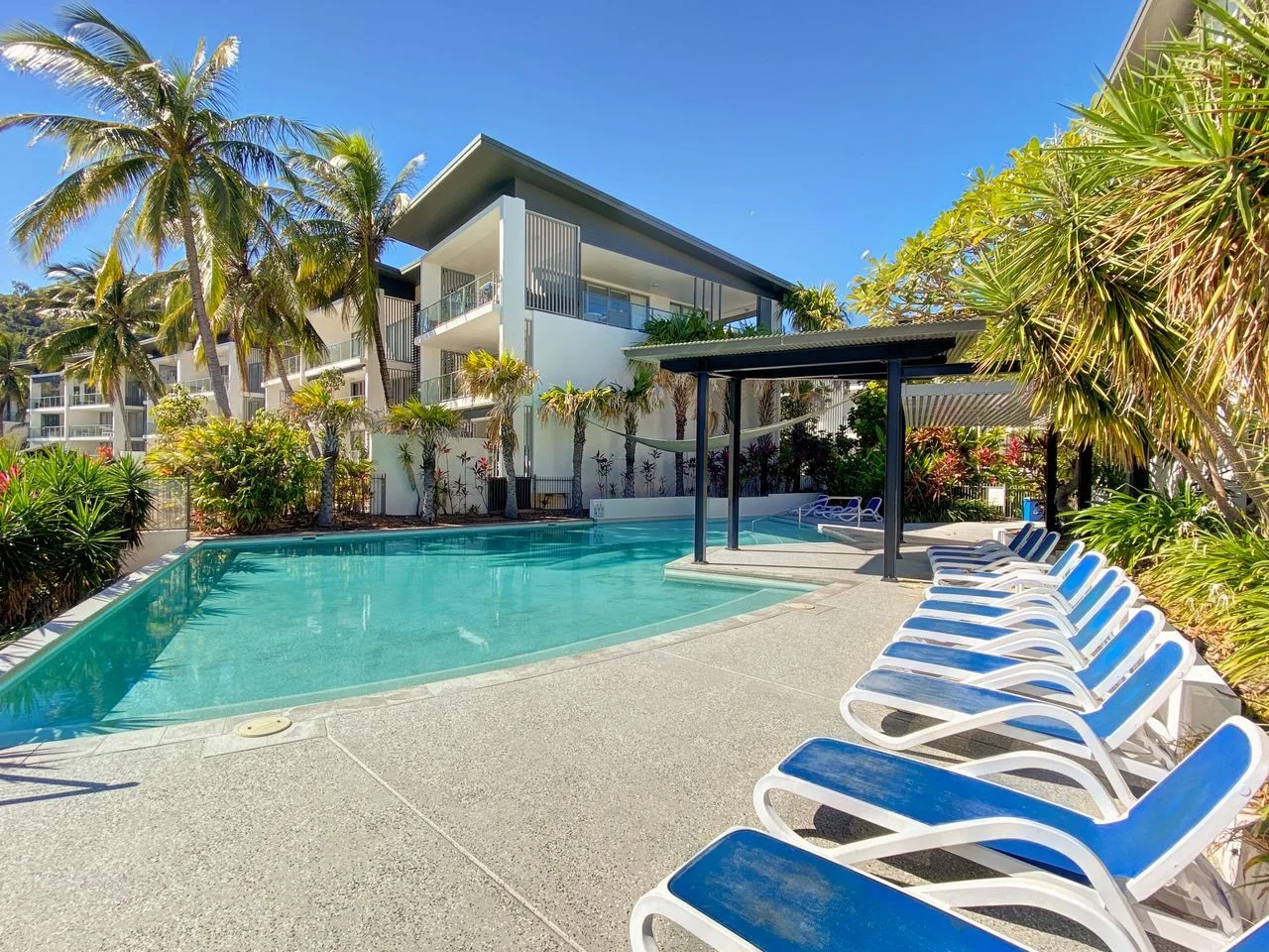 Una piscina al aire libre junto a un edificio moderno, con tumbonas azules junto a la piscina.