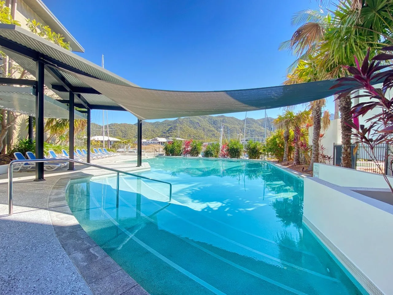 Una piscina al aire libre está bajo toldos de sombra, con palmeras y colinas al fondo.