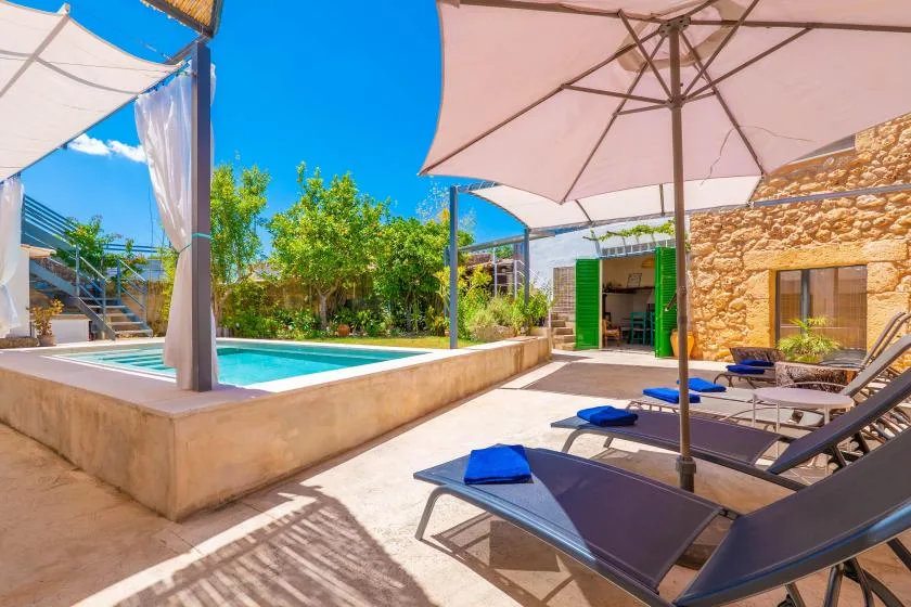 Un patio soleado con piscina, asientos reclinables y grandes sombrillas.