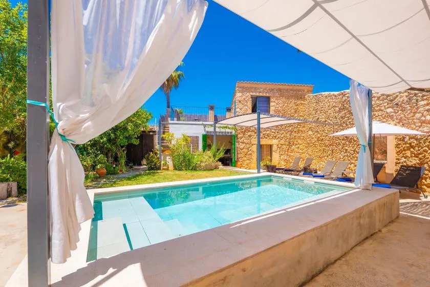 Una piscina al aire libre junto a un edificio de piedra, con asientos a la sombra cerca.
