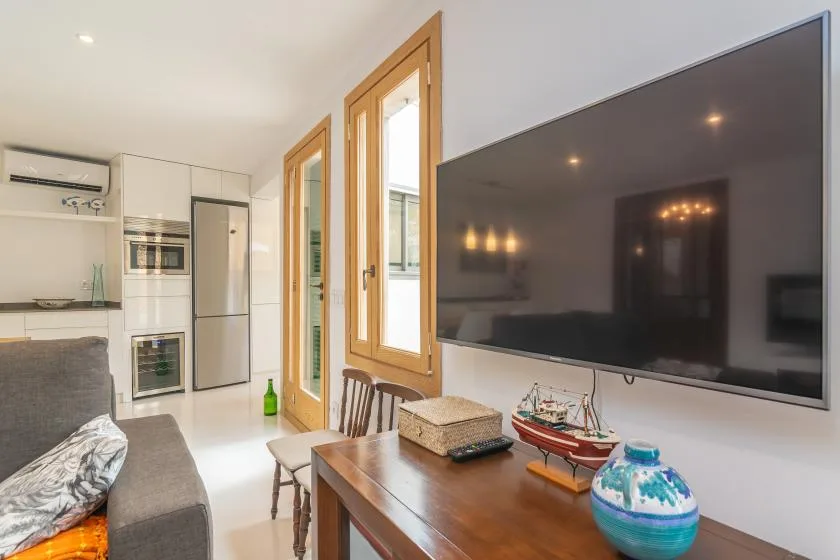 Una sala de estar con televisor, asientos y una mesa junto a ventanas altas.