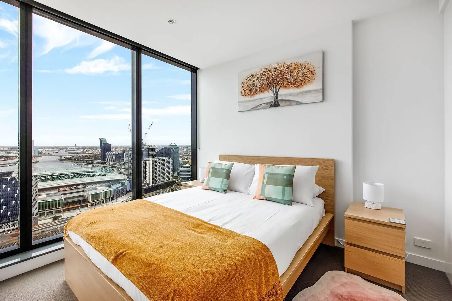 A bright bedroom has a bed beside large windows overlooking nearby city buildings.