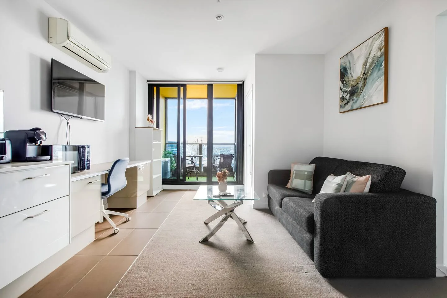 A bright living area has a dark sofa, a wall-mounted television, and glass doors opening to a balcony.