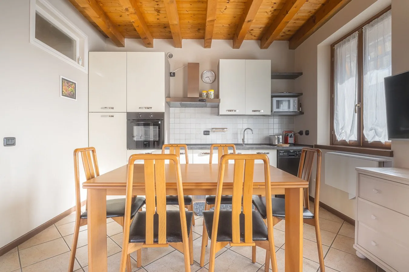 A dining table and seating sit in a bright kitchen beneath exposed wooden ceiling beams.