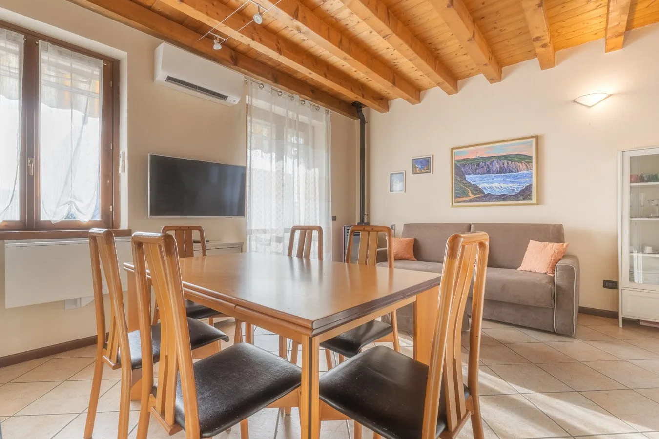 A bright living and dining area with a wooden table, seating, and a framed landscape picture.
