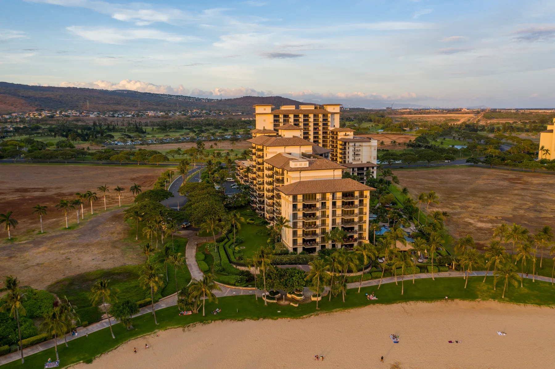 Vista aérea de un edificio turístico junto a jardines con palmeras y una playa de arena.