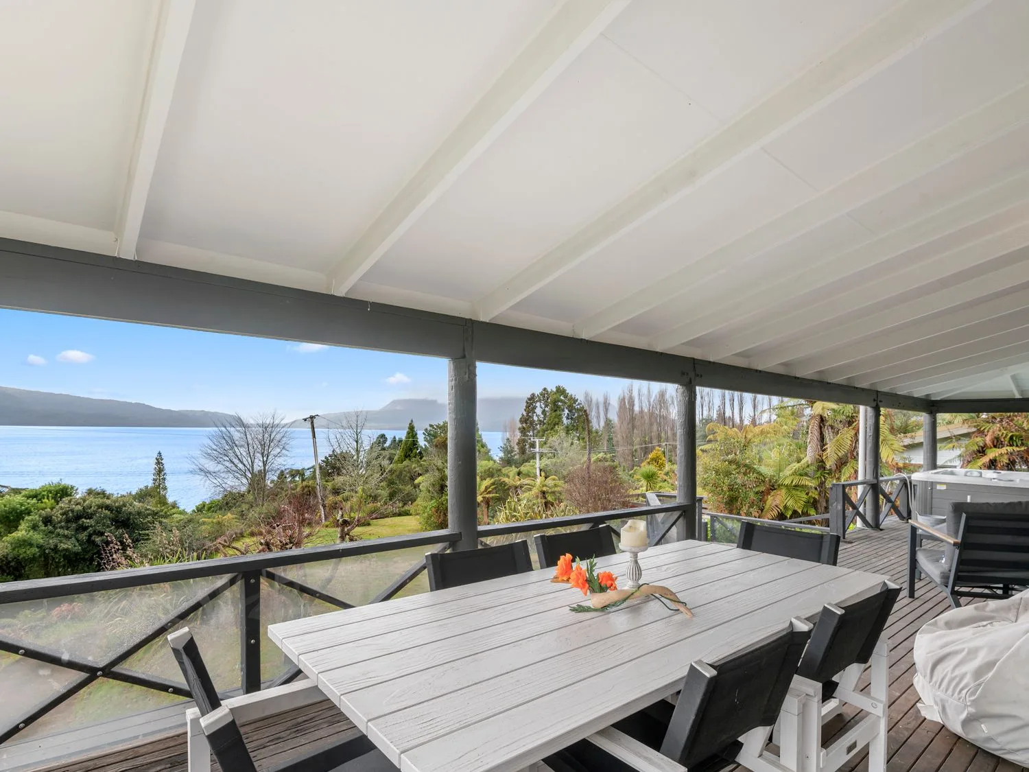 A covered wooden deck with a dining table and chairs overlooking a lake and trees at Splendour on Spencer - Lake Tarawera Holiday Home