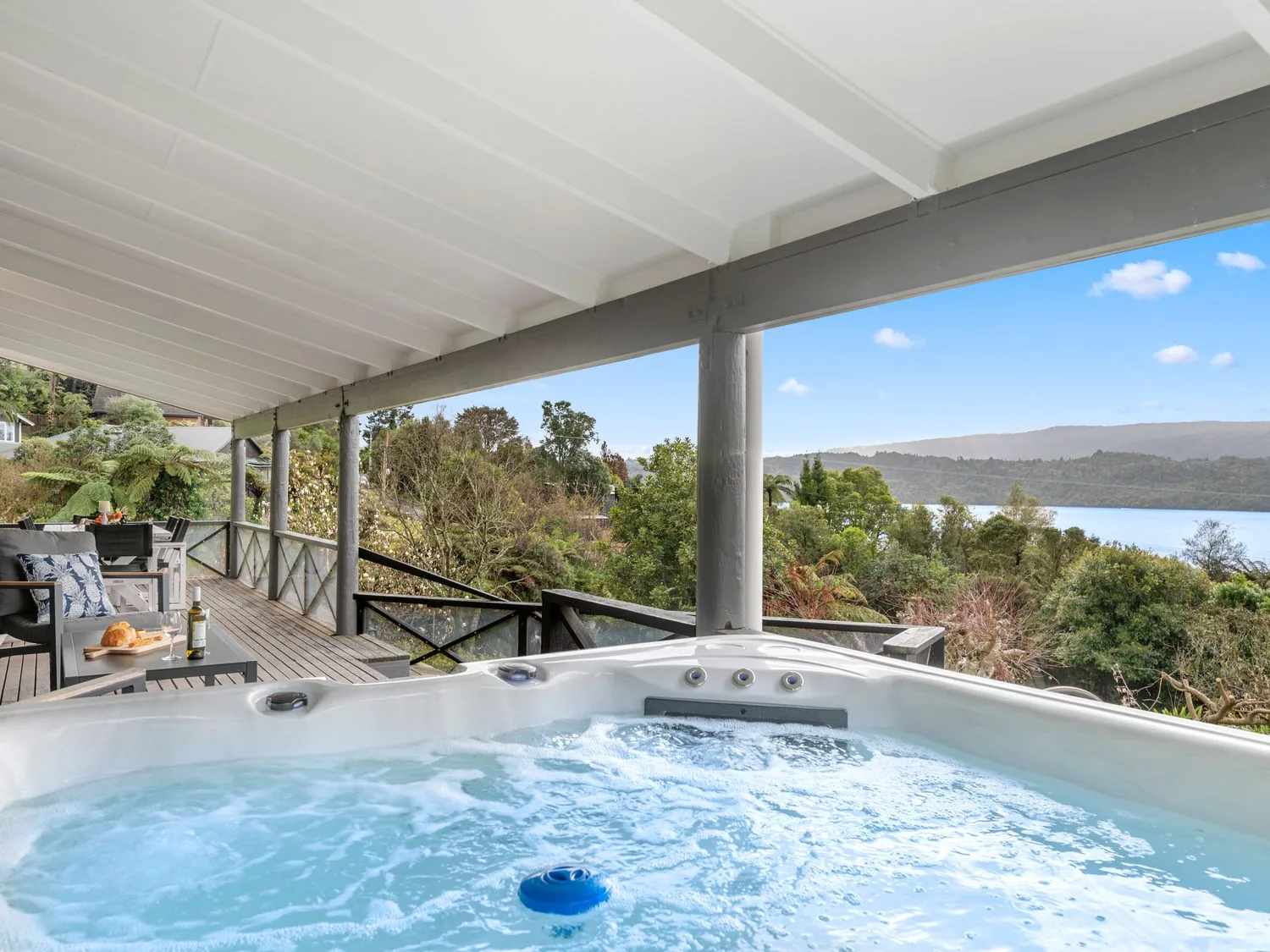 A covered wooden deck with a hot tub overlooking trees and a lake at Splendour on Spencer - Lake Tarawera Holiday Home