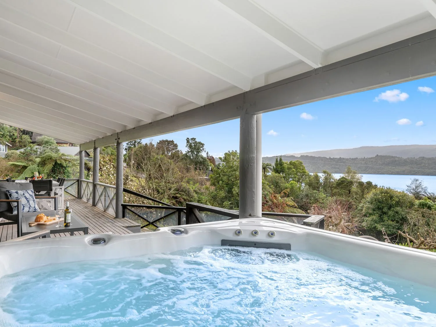 An outdoor covered deck with a hot tub a table and chairs and a view of trees and a lake at Splendour on Spencer - Lake Tarawera Holiday Home