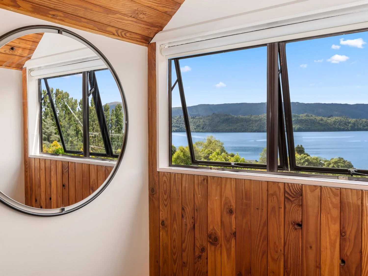 A room with wood paneling, a round mirror, and windows overlooking a lake and hills.