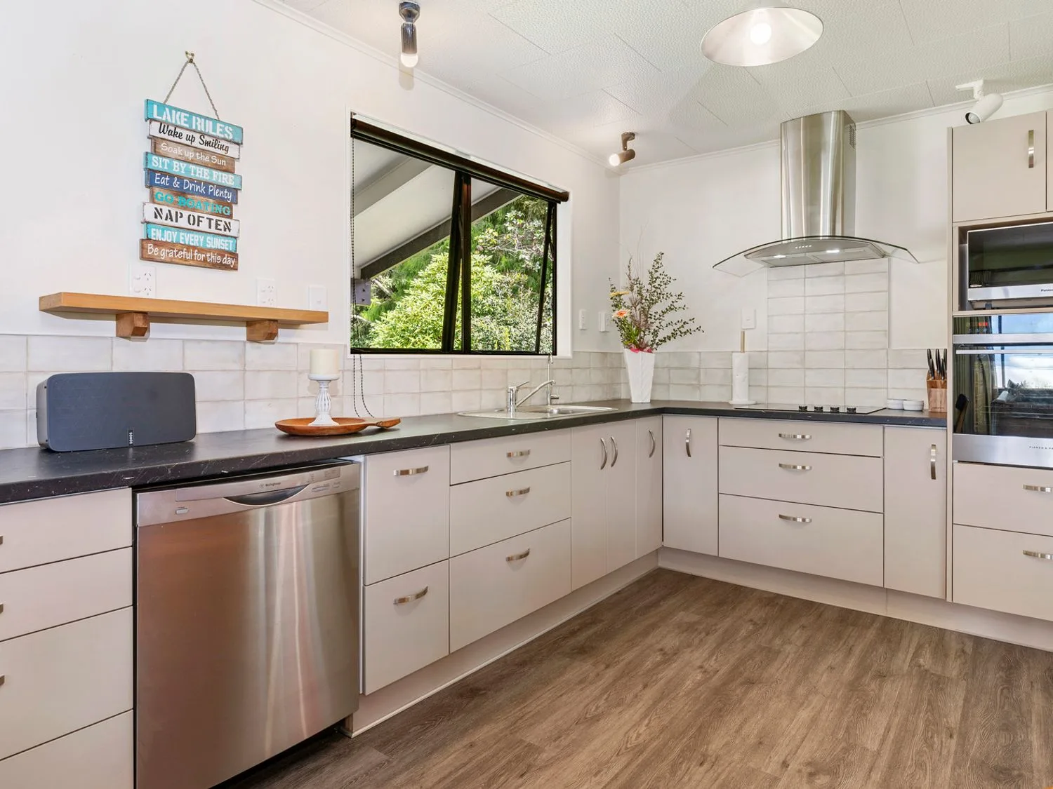 A kitchen with stainless steel dishwasher, stove, oven, sink, cabinets, window, and a decorative sign on the wall at Splendour on Spencer - Lake Tarawera