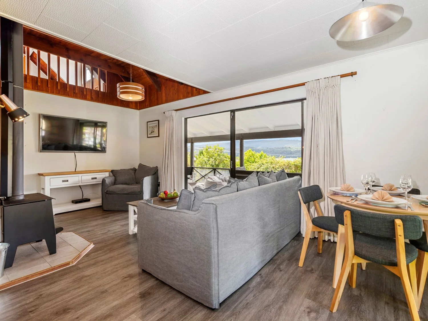 A living room with gray sofas a wood stove a wall-mounted TV and a dining area with a set table at Splendour on Spencer - Lake Tarawera Holiday Home Lake Tarawera