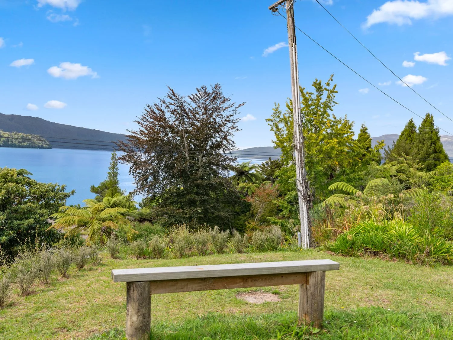 A wooden bench on grass overlooking trees water and hills at Splendour on Spencer - Lake Tarawera Holiday Home