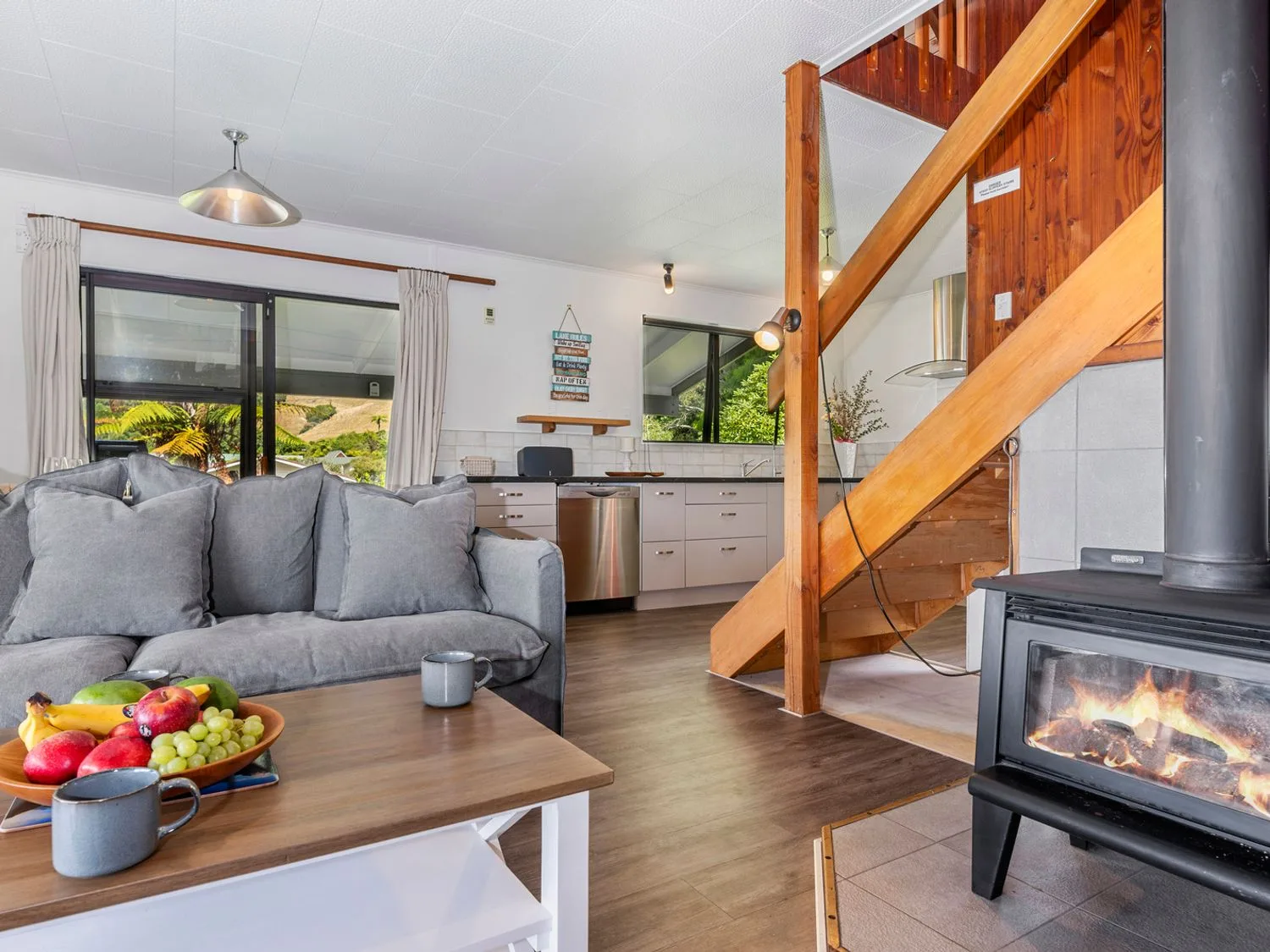 A living room with a gray sofa coffee table with fruit and mugs wood burning stove and wooden staircase at Splendour on Spencer - Lake Tarawera Holiday Home Lake Tarawera