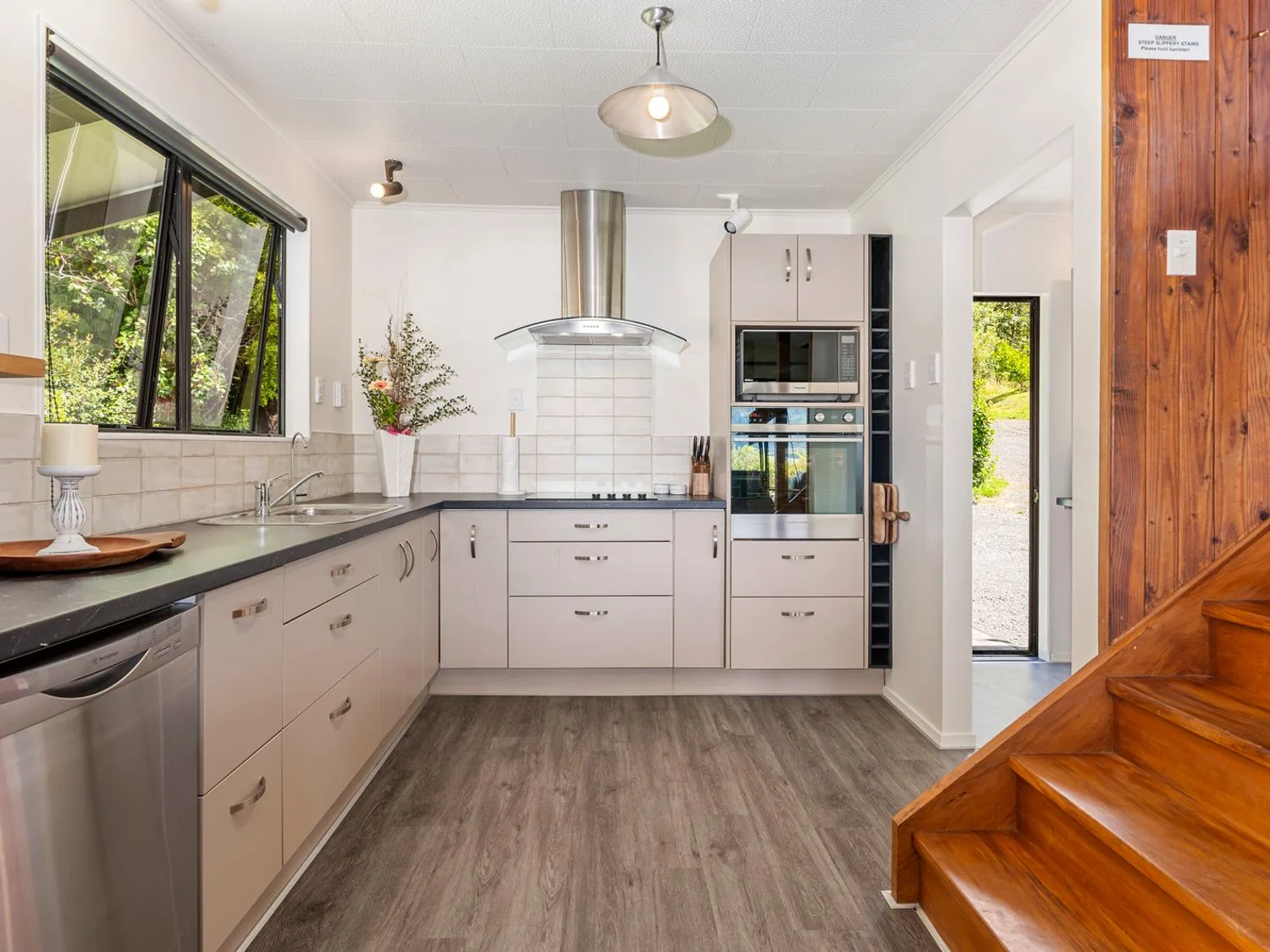 A kitchen with beige cabinets stainless steel appliances a wooden staircase and a window showing greenery at Splendour on Spencer - Lake Tarawera Holiday Home in Lake Tarawera