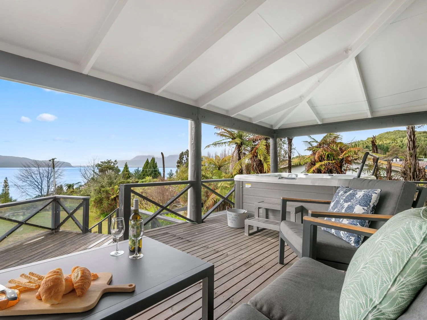 A covered wooden deck with outdoor seating a hot tub a table with wine and bread and a view of trees and water at Splendour on Spencer - Lake Tarawera Holiday Home Lake Tarawera