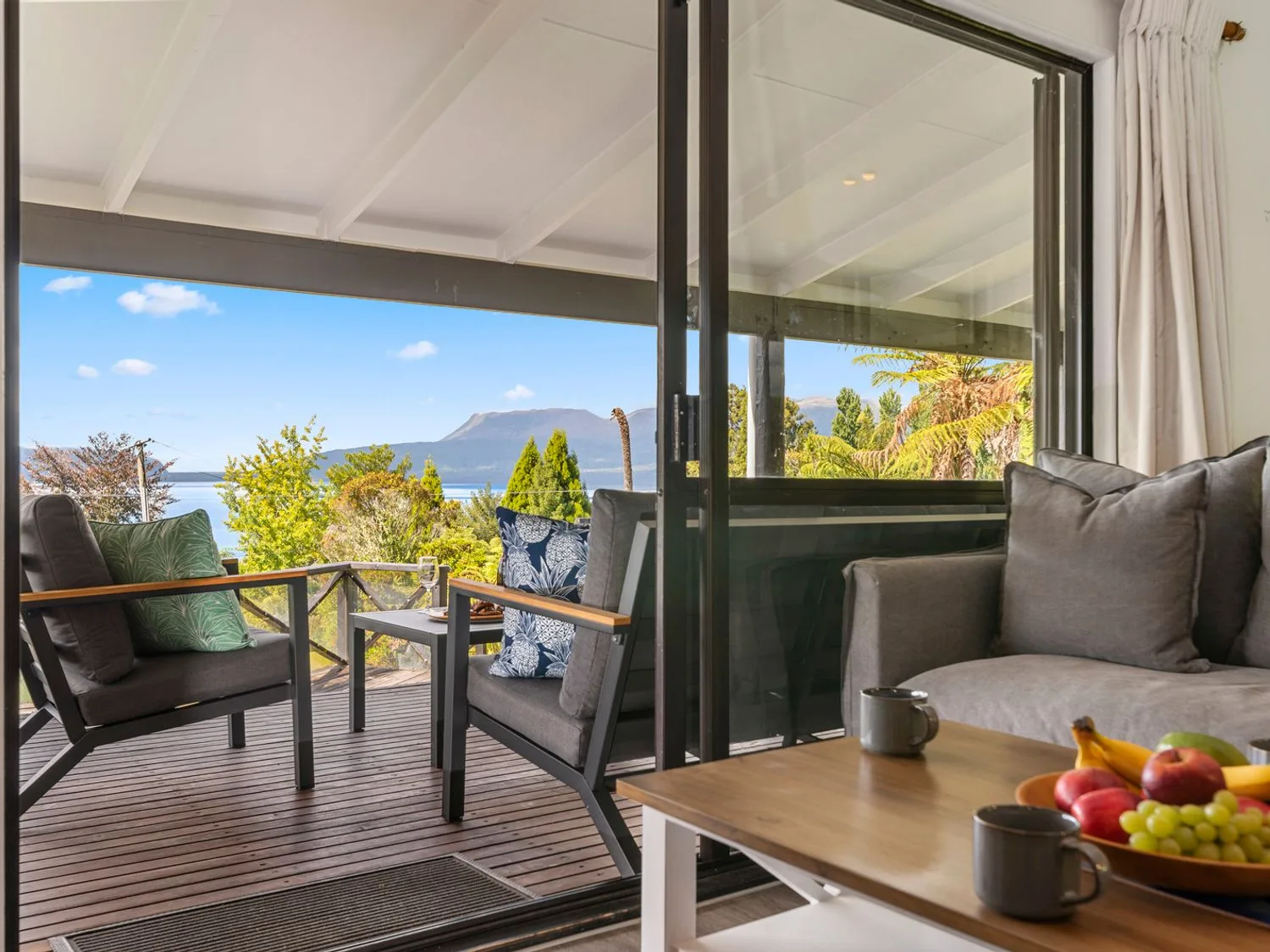 A living room opens onto a furnished deck with trees and a mountain visible beyond.