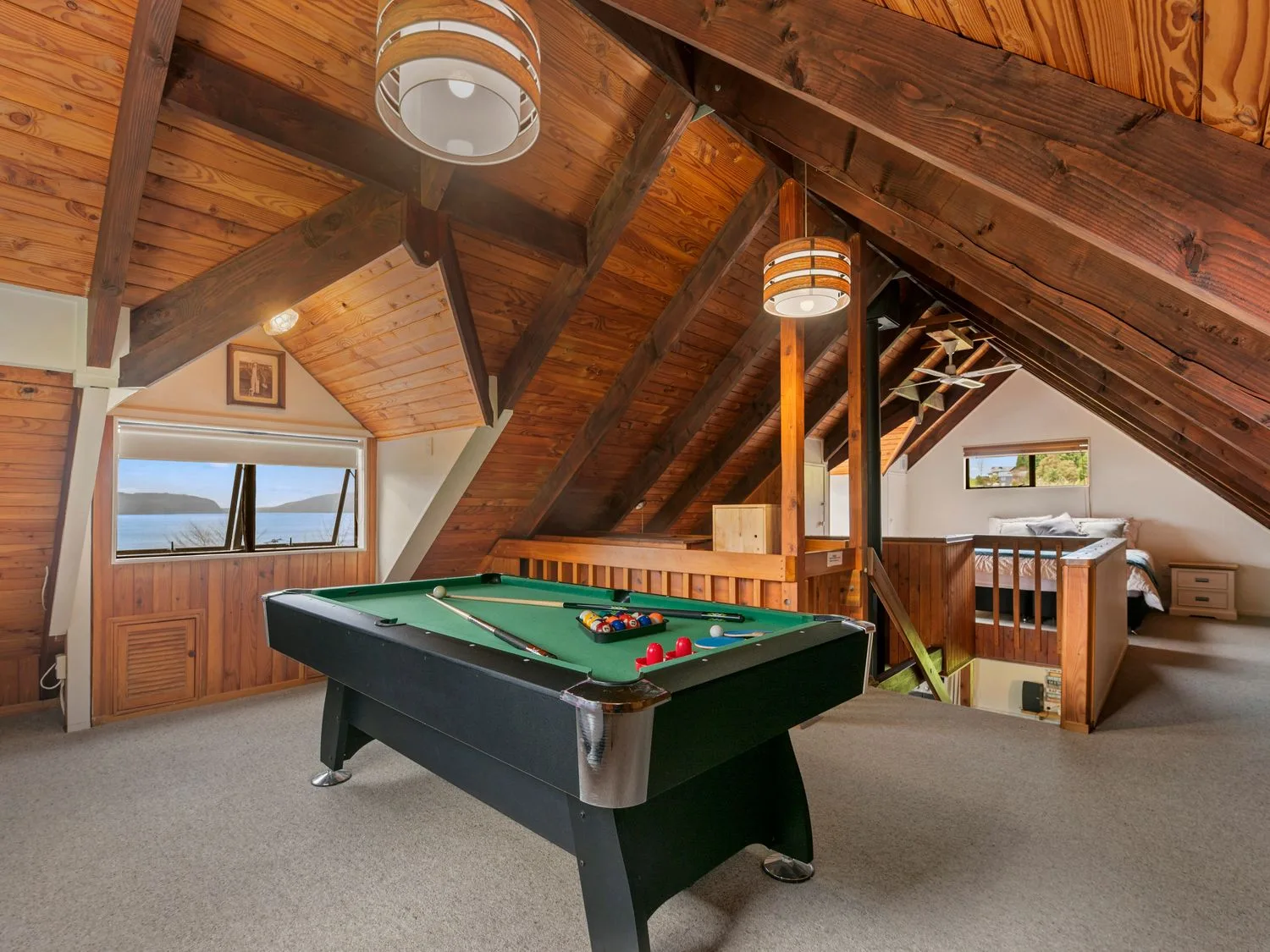 An attic room with wooden beams a pool table a bed and windows showing a lake view at Splendour on Spencer - Lake Tarawera Holiday Home